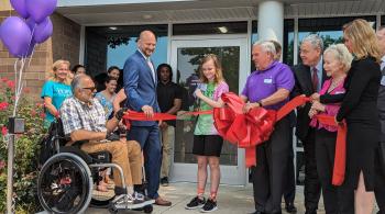 A group of at least 16 people of different ages gathers outside a glassed-in entrance with “Kennedy Krieger” above the doors. The group is holding a long, wide red ribbon with a giant bow in the center. A young person wearing a shirt that says “HOPE” is cutting the ribbon with giant scissors. Holding one end of the ribbon is a man sitting in a wheelchair. Some people are wearing suits, while others are wearing more casual clothing.