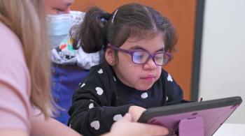 A young girl, assisted by a therapist, uses a tablet-like device that helps her communicate.