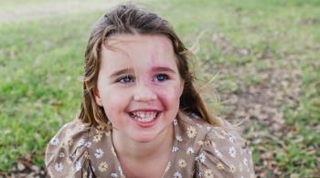 Rainey leans forward and smiles at the camera while sitting on the ground outside. 