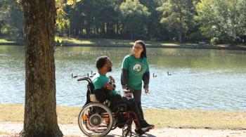 A man and woman talk on a pathway that winds around a pond. The man is sitting in a wheelchair, while the woman is standing to his left looking at him.