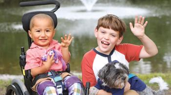 Posed photo of a young boy sitting in a wheelchair and a slightly older boy holding a small dog. Both boys are smiling and waving.