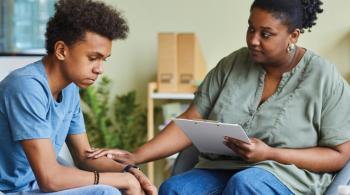 A woman holding a clipboard and with a concerned expression on her face places one of her hands on the arm of a teenage boy.