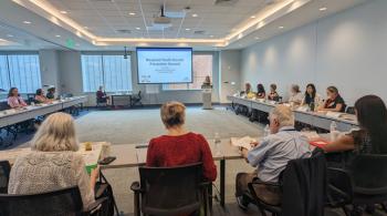 A woman speaks in front of a projection screen, which displays a slide that says Maryland Youth Suicide Prevention Summit. She is speaking to a group of attendees inside a conference room.