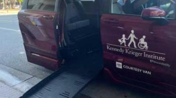 A wheel-chair accessible Toyota Sienna is parked at the curb with the ramp lowered. The adaptive van is burgundy and has Kennedy Krieger Institute's logo on the front passenger door. 