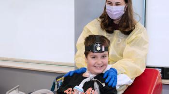 Posed photo of a smiling boy with a woman behind him. The woman is wearing a yellow medical gown and a face mask and has her hands on the boy’s shoulders. Her squinting eyes indicate she is smiling under her face mask. The boy is smiling and wearing a shirt that says “Orioles” on it. He has clear plastic tubes attached to his neck for breathing purposes and is wearing a head lamp-like device. Two electrodes marked “Leo” are attached to his left arm.