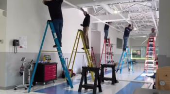 Five adults stand atop colorful ladders in an open interior space while adjusting ceiling apparatus.