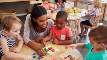 A teacher smiles while helping three young students with an activity involving wooden shapes. Three other students are seen completing a similar activity in the background. 