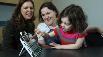 A young girl completes an activity using a tablet, while two women over her right shoulder smile.