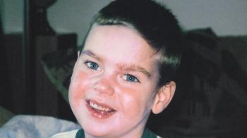 A young boy with dark blue-green eyes and blonde hair smiles for the photo. He is wearing a white T-shirt with dark green edging.