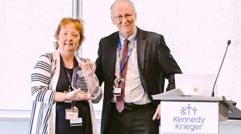 Dr. Peter van Zijl and a Kirby Center staff member stand next to a podium during the 25th anniversary symposium. The staff member is holding an award in her hands.