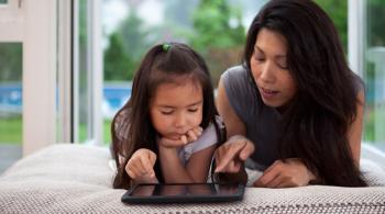 A mother and her young daughter lay across a sofa while looking at a tablet.