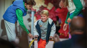 Un niño pasea por la pasarela de Fall Fête 5 usando un dispositivo de movilidad. Un chico y una mujer se colocan a ambos lados para ofrecerle ayuda.