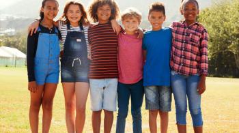 Six pre-teen friends stand shoulder to shoulder while smiling in a park.