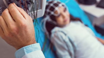 Close up of a laboratory worker's hand setting up an electroencephalography machine before analyzing brain of a little girl lying on an examination couch in the background.
