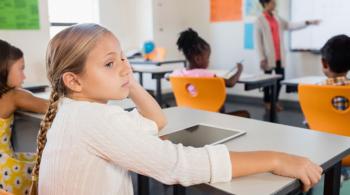 A little girl gazes off to the side as she sits in a classroom. She is looking away from her teacher, who is writing on a whiteboard as the other students watch.