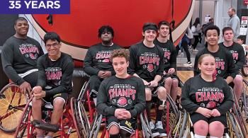 Nine teens pose for the camera. Each sits in a wheelchair, and they appear to be on a basketball court. Each teen wears a black T-shirt that says “Conference Champs” on it.