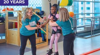 A smiling young girl is suspended in a harness from a weight-supporting device. On her feet are black braces that support her feet and ankles. Helping her are two women, both wearing dark pants and teal T-shirts that say “Team Kennedy Krieger.” In the background are colorful therapy balls and other physical therapy equipment.