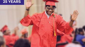 A young man in a red graduation cap and gown walks by a group of cheering people, including other young people in red graduation caps and gowns. The young man is smiling and looking down, and he has his hands up in the air.