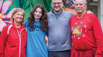 Four people smile for the camera with their arms around each other’s backs. On the far left and far right are a woman and a man in red athletic clothing. Right of center is a man with a gray shirt with the Kennedy Krieger logo on it. Left of center is a young woman wearing a blue T-shirt that says “ROAR for Kids.”