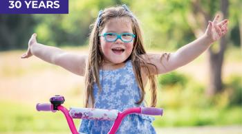 A smiling young girl wearing aqua-framed glasses sits on a purple and pink bike outside, her arms stretched widely. The bike is stationary.