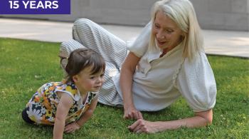 A very young girl crawls on the grass outside, while WIN co-president Cynthia Cavanaugh smiles beside her. Cavanaugh appears to be encouraging the girl to crawl.
