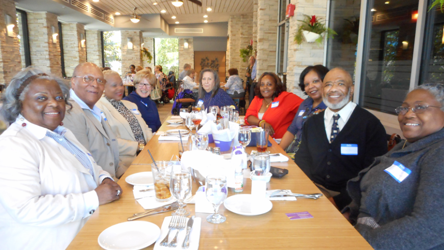 A group of nine, six women and three men, sit along a rectangular table. 