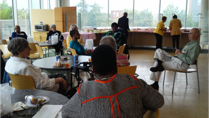 A man sitting in a chair speaks as alumni members look on from their tables.