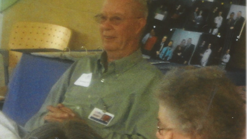 A man in a green shirt sits and smiles. He is facing to the left of the camera.