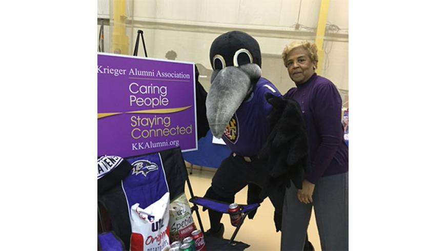 A woman and Poe, the Baltimore Ravens mascot, stand next to the Kennedy Krieger Alumni table. 