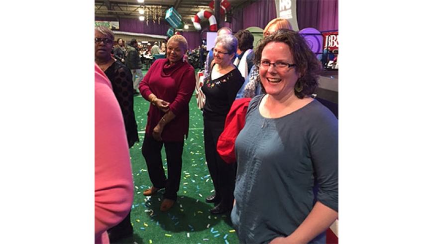 A smiling woman in a gray short stands to the right with other women in the background.