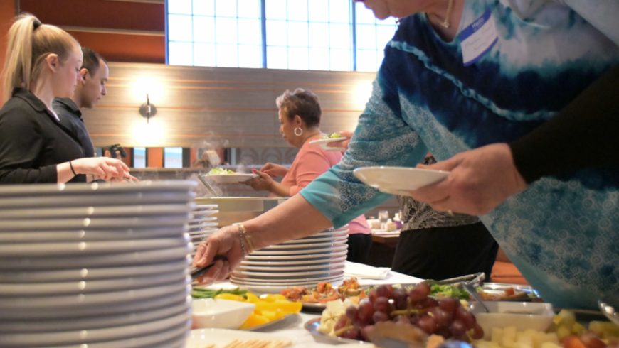 Event attendees take food and plates from buffet table. 