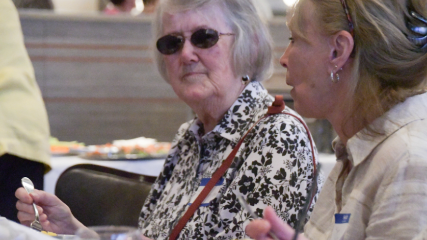 A women sitting at a table while wearing dark glasses faces the camera, with another woman sitting off to her left.