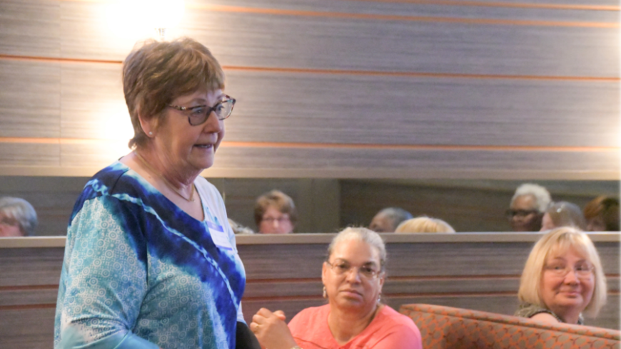 A woman stands and speaks, with seated attendees in the background. 