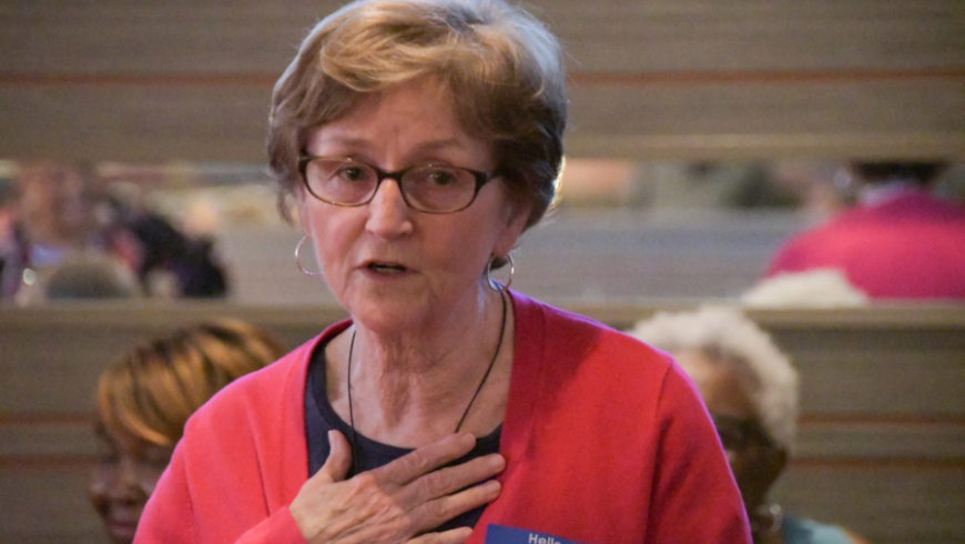 A woman in a red shirt holds her right hand to her chest while speaking.