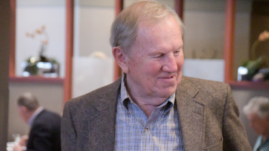 A man in a gray jacket and collared shirt looks to his right.