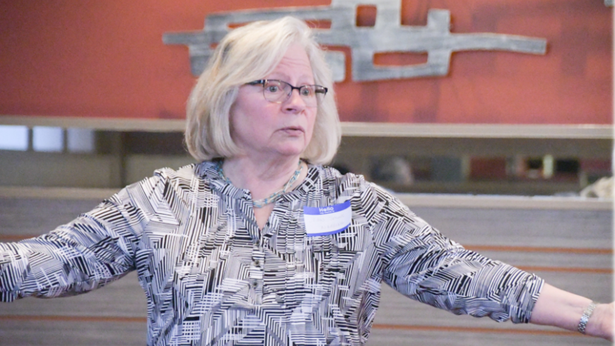 A woman in a gray shirt stands and speaks with her arms out to her side. 