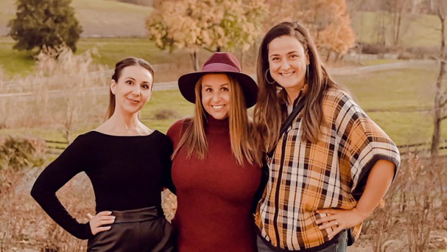 Three women pose for a photo outside, with a hill, trees, and a wooden fence behind them. 