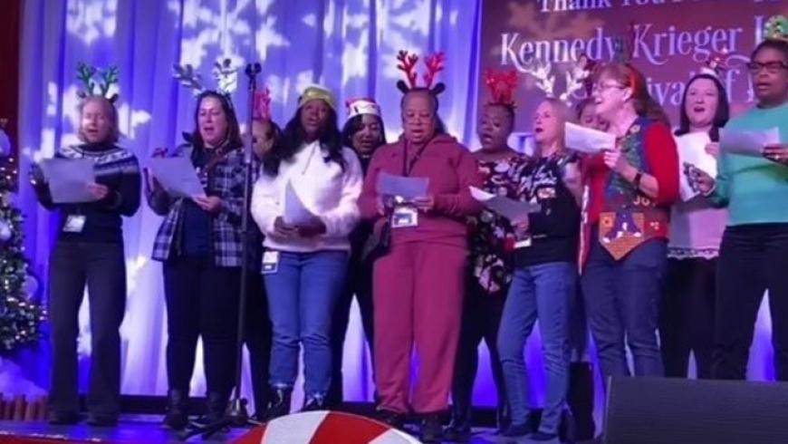 Unit Clerks, Nursing Technicians, RNS, Care Managers, Nurse Educators, and Nursing Leadership sing on stage at Festival of Trees. 