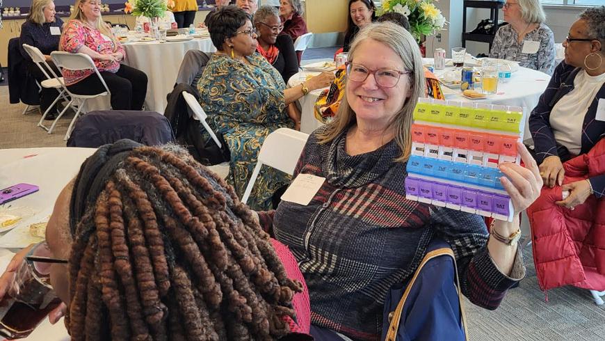A woman sits at a table and smiles while holding up a colorful piece of paper. Another person is sitting with their back to the camera, while others are seen sitting at adjacent tables.