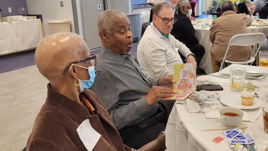 Three people sit at a table and talk. One of them is wearing a protective facemask.