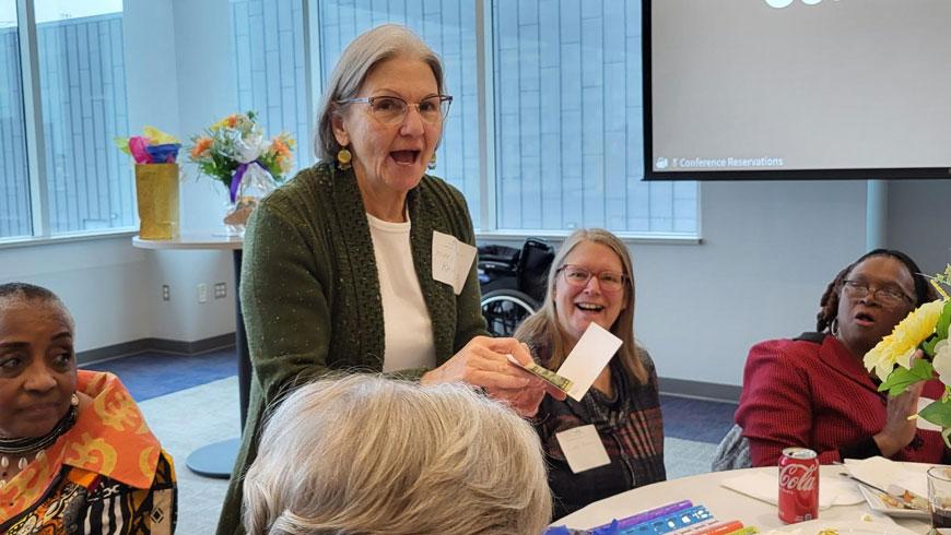 A woman stands and speaks to the room from her table.