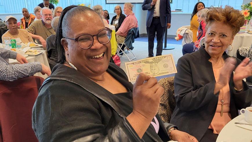 A woman smiles while holding a $50 bill upside down. The woman seated next to her is smiling and clapping.