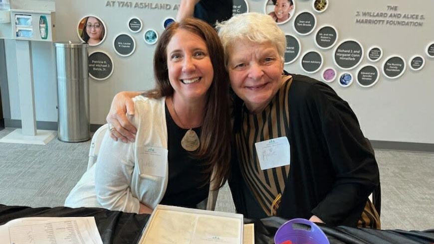 Two women wrap their arms around each other's shoulders and smile while sitting next to each other.