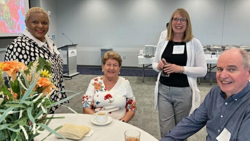 Three women and a man smile for a photo. Two of the women are standing to either side of a woman who is sitting, while the man is sitting next to her. 