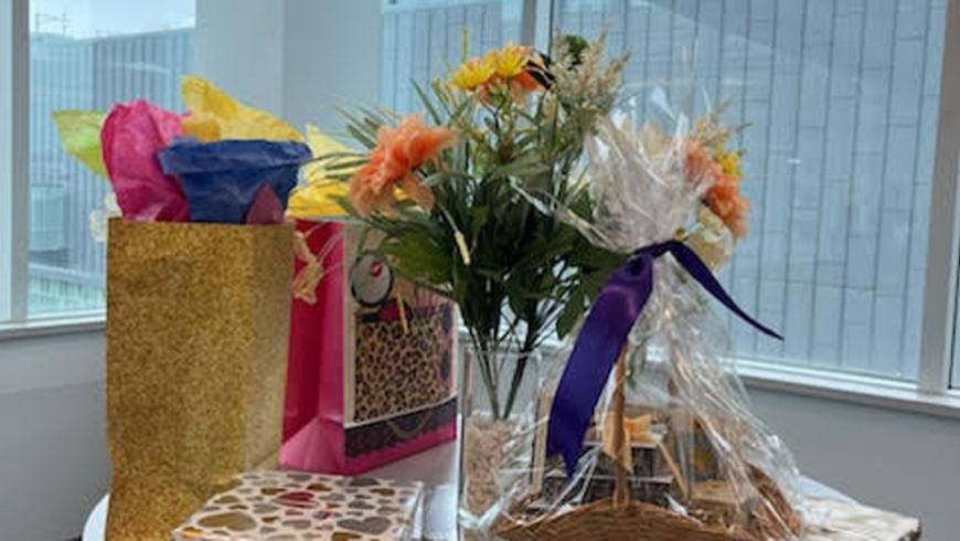 A glass vase while flowers sits on a table. It is surrounded to one side by side by a wrapped present and two gift bags. A gift basket sits off to the other side.