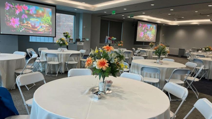 An empty conference room is staged for the event. Banquet tables with white table cloths and flowers in vases as centerpieces can be seen in the foreground, with two projection screens in the background.