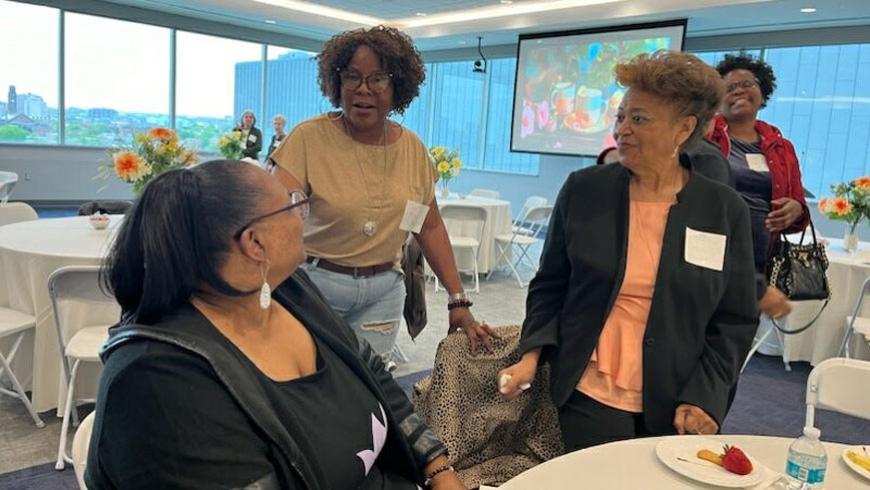 A woman sitting at a table looks over shoulder to talk to two women standing behind her.