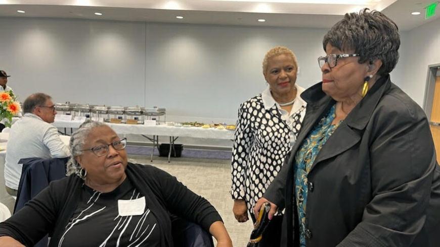 A woman sitting at a table looks to one side to talk to a woman standing next to her. Another woman is seen walking towards them.