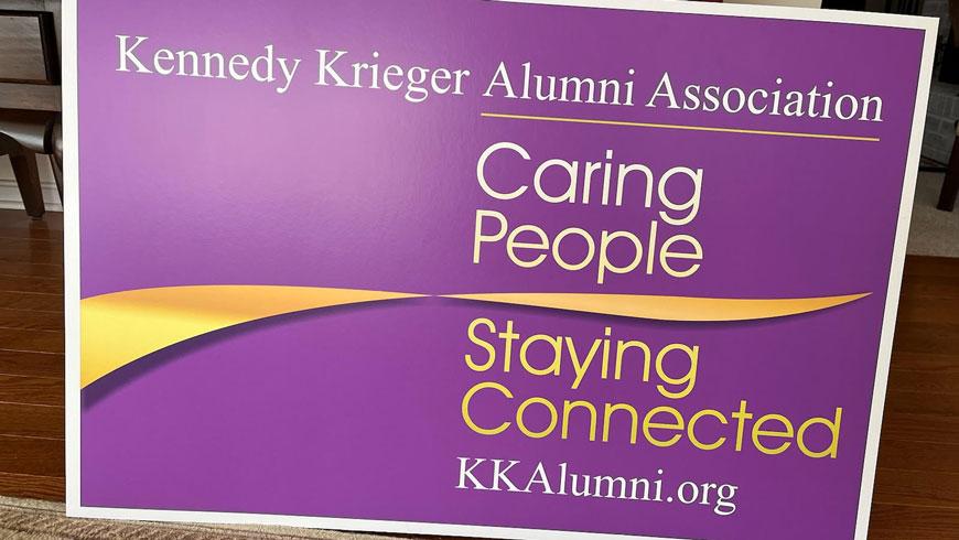 A purple sign with the Kennedy Krieger Alumni Association's logo. Below the association's name is text that reads Caring People Staying Connected KKAlumni.org. A gold swirl crosses the sign between the words People and Staying