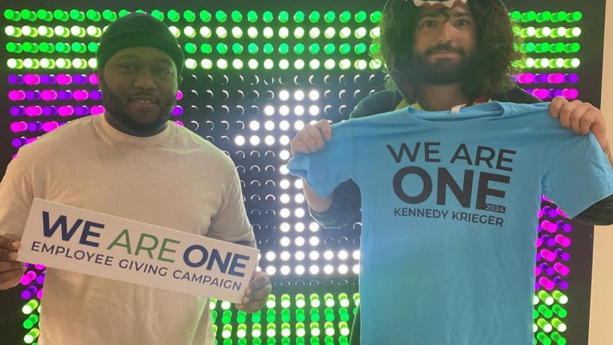 Two staff members standing in front of a brightly lit Kennedy Krieger Schools sign. One of them is holding a We are One sign, while the other is wearing a We Are One t-shirt.
